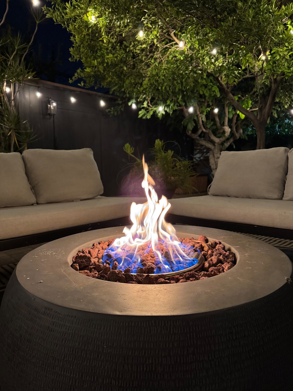 The fire pit burning at night, ringed by outdoor sofas under string lit trees